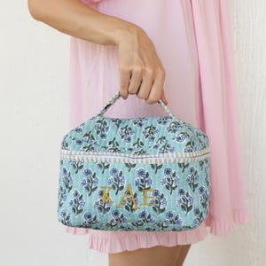 Pode incluir: Uma bolsa de cosméticos acolchoada com um padrão floral turquesa e as letras "KATE" bordadas em dourado. A bolsa tem uma alça e uma borda listrada. A bolsa é segurada por uma pessoa vestindo um vestido rosa.
