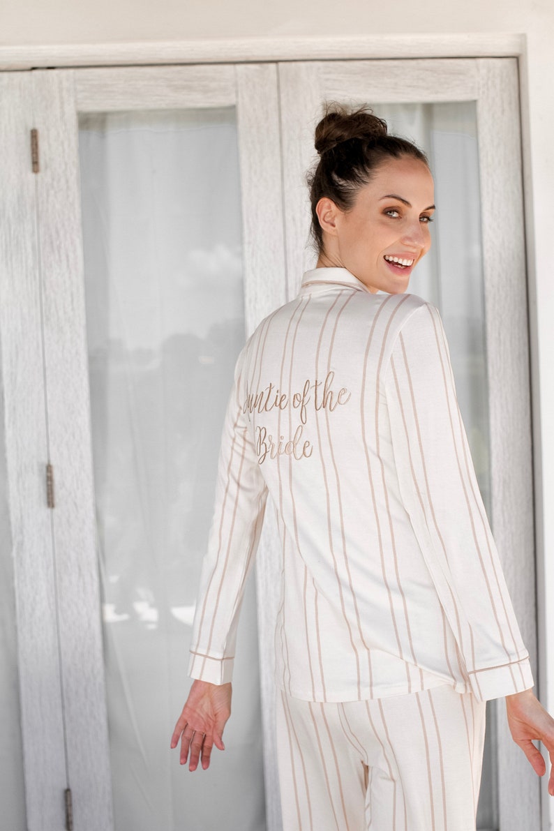 Puede incluir: Una mujer con un pijama de rayas blancas y marrones con el texto "Auntie of the Bride" bordado en la espalda.