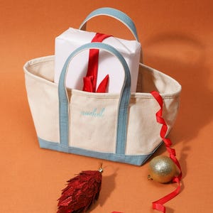 May include: A cream canvas tote bag with light blue trim and handles, embroidered with the name "rachel". A wrapped gift with a red ribbon is inside the bag, with a red ribbon, a gold bauble, and a red pinecone ornament nearby.