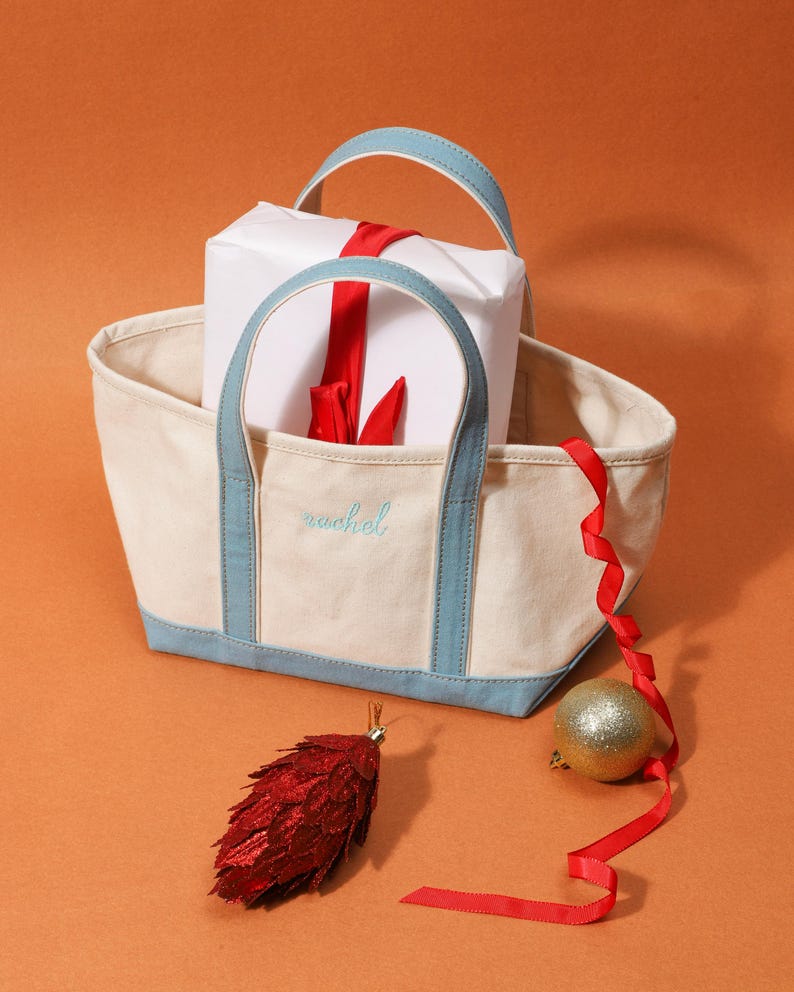 May include: A cream canvas tote bag with light blue trim and handles, embroidered with the name "rachel". A wrapped gift with a red ribbon is inside the bag, with a red ribbon, a gold bauble, and a red pinecone ornament nearby.