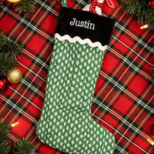 May include: A green Christmas stocking with a white tree pattern and a black cuff embroidered with the name "Justin" in white. A small polar bear wearing a red scarf peeks out from the top. The stocking is set against a red and green plaid background.