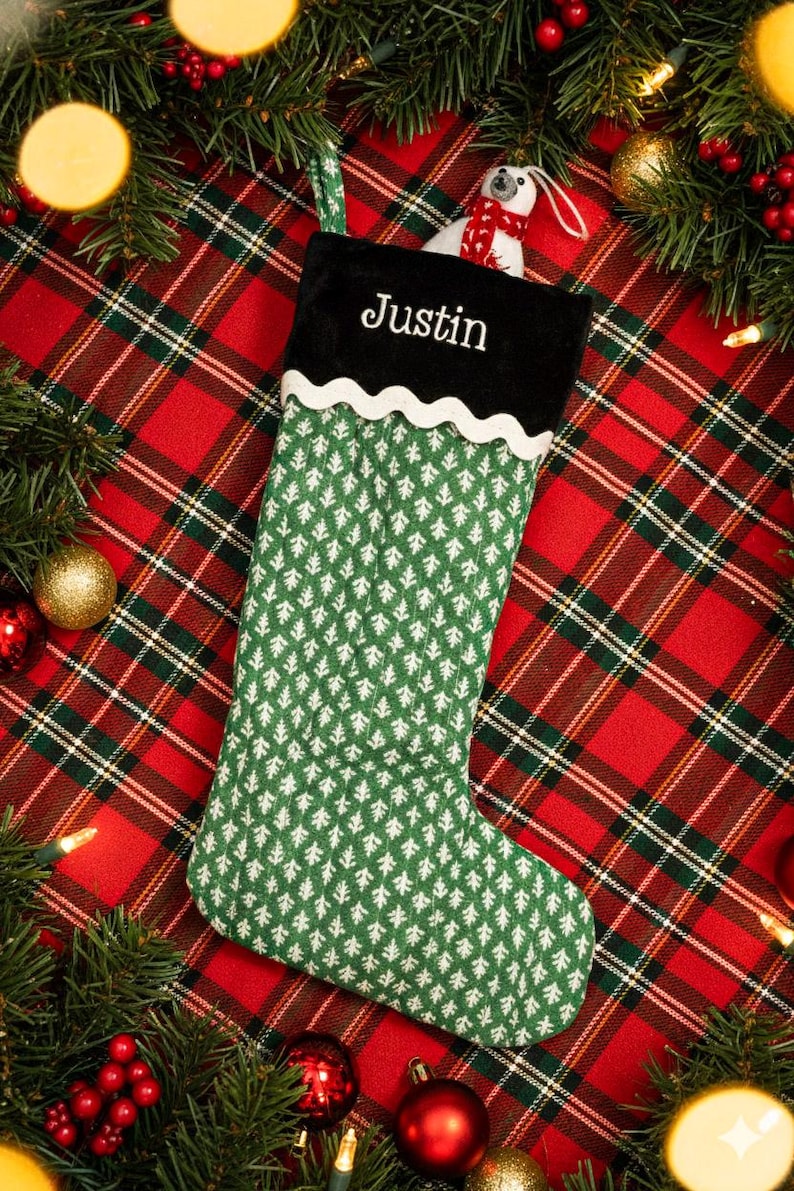 May include: A green Christmas stocking with a white tree pattern and a black cuff embroidered with the name "Justin" in white. A small polar bear wearing a red scarf peeks out from the top. The stocking is set against a red and green plaid background.