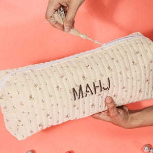 May include: Cream-colored, quilted pencil case with a floral pattern and the embroidered letters "MAHJ." The rectangular case has a white zipper and is being held open by a hand. The tassel is held by another hand.