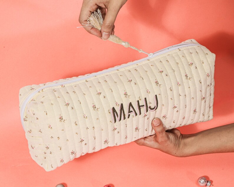 May include: Cream-colored, quilted pencil case with a floral pattern and the embroidered letters "MAHJ." The rectangular case has a white zipper and is being held open by a hand. The tassel is held by another hand.