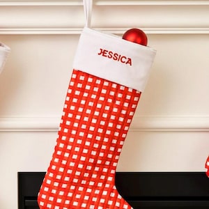 May include: Red and white checkered Christmas stocking with the name "JESSICA" embroidered in red. A red ornament sits inside the stocking. The stocking hangs from a white mantel decorated with greenery, red berries, and pinecones.