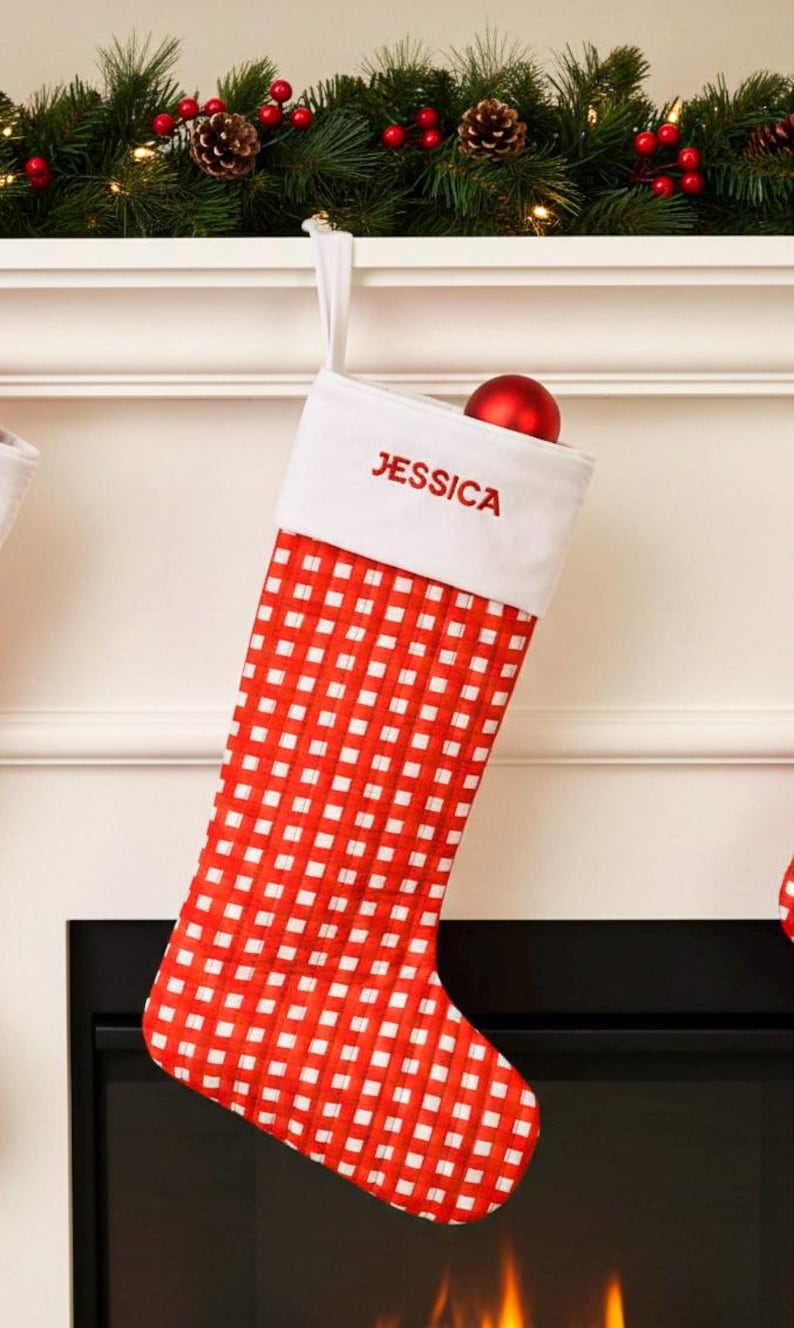 May include: Red and white checkered Christmas stocking with the name "JESSICA" embroidered in red. A red ornament sits inside the stocking. The stocking hangs from a white mantel decorated with greenery, red berries, and pinecones.