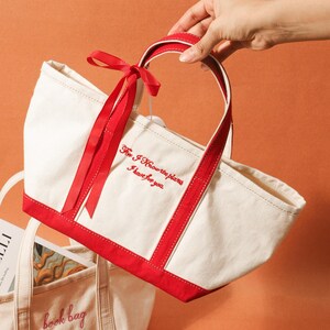 May include: A cream canvas tote bag with red edging and handles. The bag has the embroidered text "For I Know the plans I have for you" in red. A red ribbon is tied to the handle. The bag is held by a person.