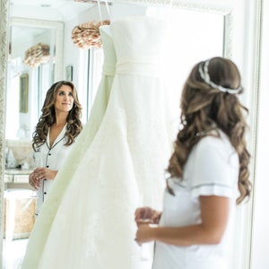 May include: A bride-to-be in a white pyjama top and shorts looks at her wedding dress hanging in a mirror.