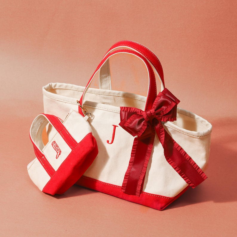 May include: A canvas tote bag and a smaller matching bag. The bags are off-white with red trim and handles. A large red bow adorns the larger bag. The letter "J" is embroidered on the front of the larger bag. The smaller bag has a red embroidered design.
