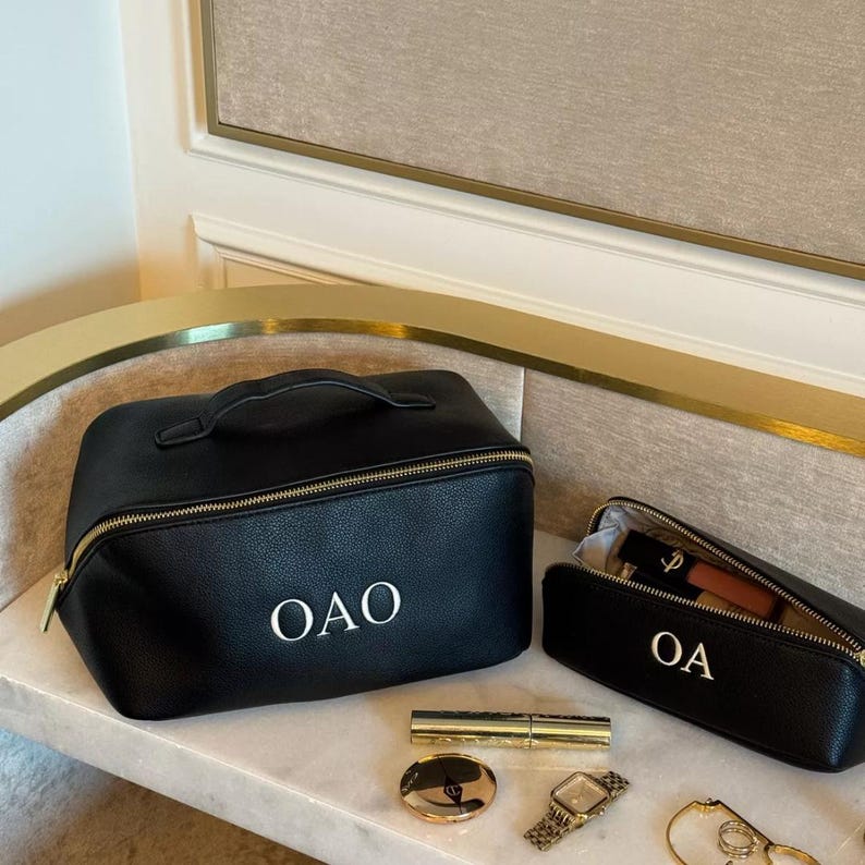 May include: Two black leather zippered travel makeup bags with gold zippers. The larger bag has a handle and the initials "OAO" embroidered in white. The smaller bag has the initials "OA" embroidered in white.