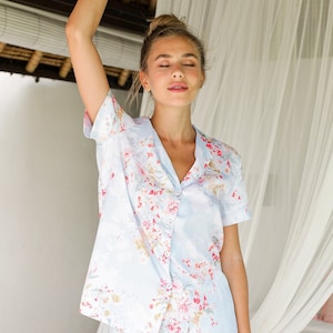 Puede incluir: Una blusa de pijama azul claro con estampado floral. La blusa tiene mangas cortas y cierre de botones en la parte delantera.