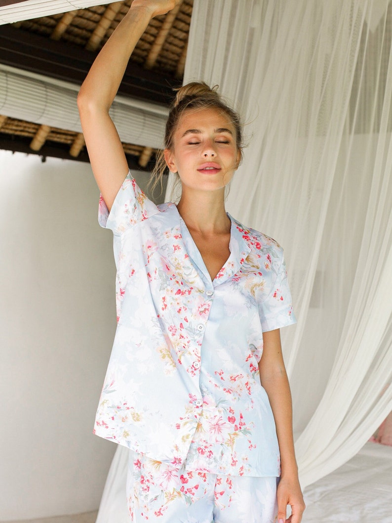 Puede incluir: Una blusa de pijama azul claro con estampado floral. La blusa tiene mangas cortas y cierre de botones en la parte delantera.