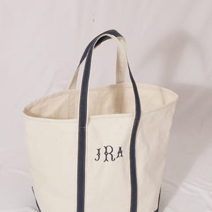 May include: Cream-colored canvas tote bag with navy blue accents. The bag features navy blue handles and trim, and the initials "JRA" are embroidered in navy blue on the front. A simple, classic design.