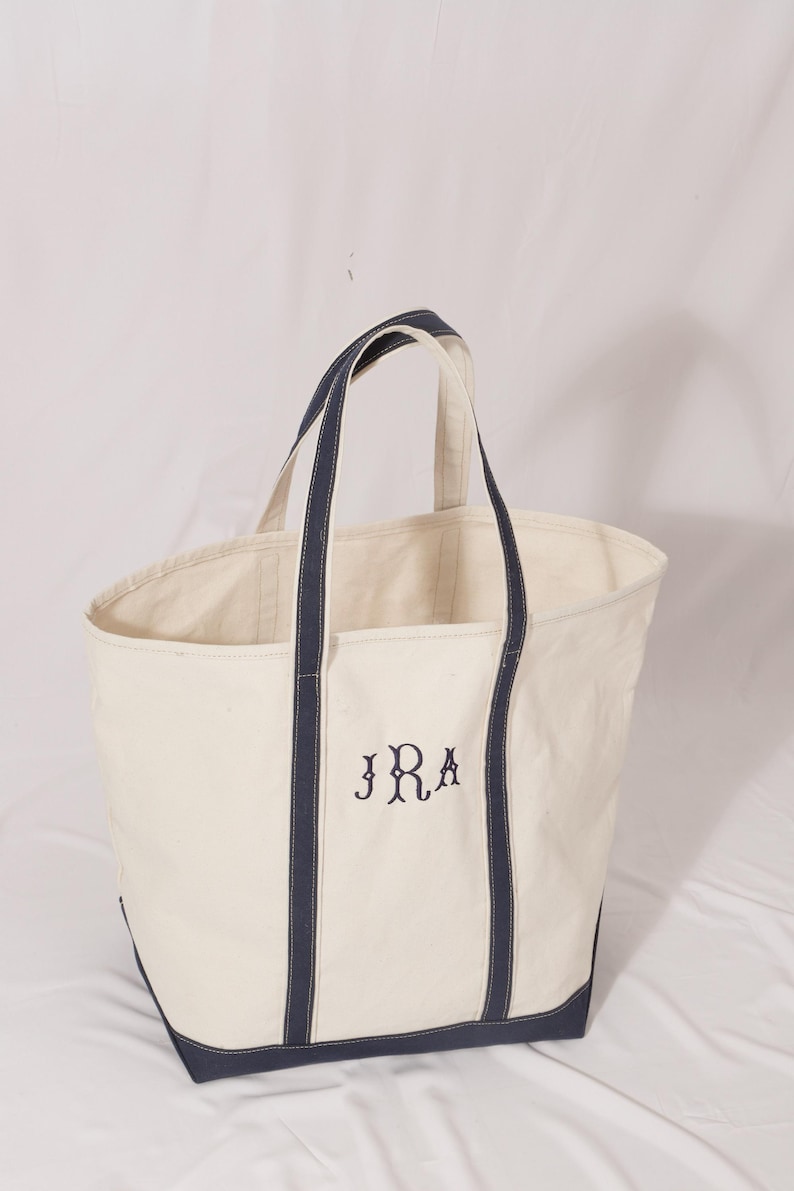 May include: Cream-colored canvas tote bag with navy blue accents. The bag features navy blue handles and trim, and the initials "JRA" are embroidered in navy blue on the front. A simple, classic design.