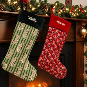 May include: Two Christmas stockings hang above a fireplace. One is green with a leopard print and the name "Ashley" embroidered on the cuff. The other is red with a floral pattern and the name "Michelle". Festive garland and lights decorate the mantel.