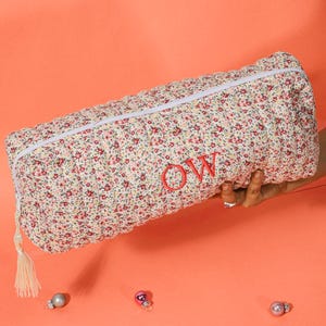 May include: A floral print pencil case with a white zipper and tassel. The case is cream-colored with a pattern of small red and pink flowers. The initials "OW" are embroidered in red on the case.