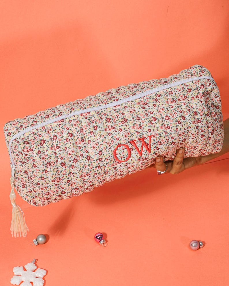 May include: A floral print pencil case with a white zipper and tassel. The case is cream-colored with a pattern of small red and pink flowers. The initials "OW" are embroidered in red on the case.