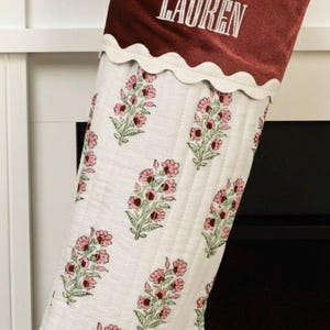 May include: A Christmas stocking with a burgundy velvet cuff embroidered with the name "LAUREN" in white. The body of the stocking is white with a floral pattern in pink and green. A scalloped white trim separates the cuff from the body.