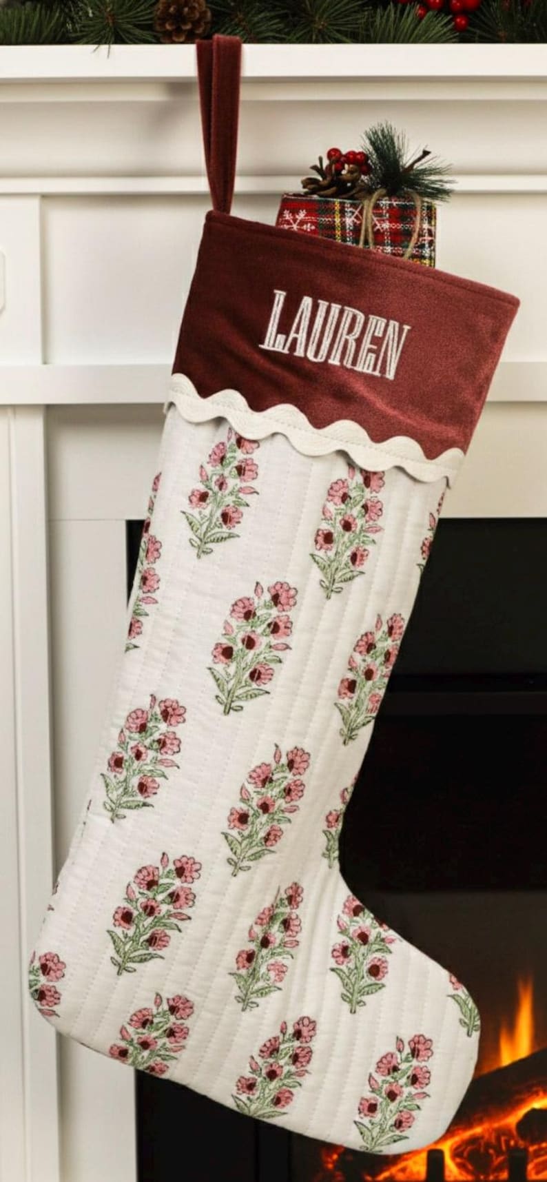 May include: A Christmas stocking with a burgundy velvet cuff embroidered with the name "LAUREN" in white. The body of the stocking is white with a floral pattern in pink and green. A scalloped white trim separates the cuff from the body.