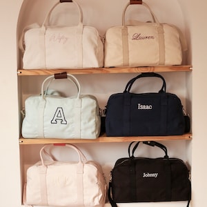 May include: Five canvas duffel bags with personalized embroidery on a wooden shelf. The bags are in various colors, including beige, light blue, navy blue, and black. The embroidery includes the names "Wifey", "Lauren", "Isaac", "ACB", and "Johnny".