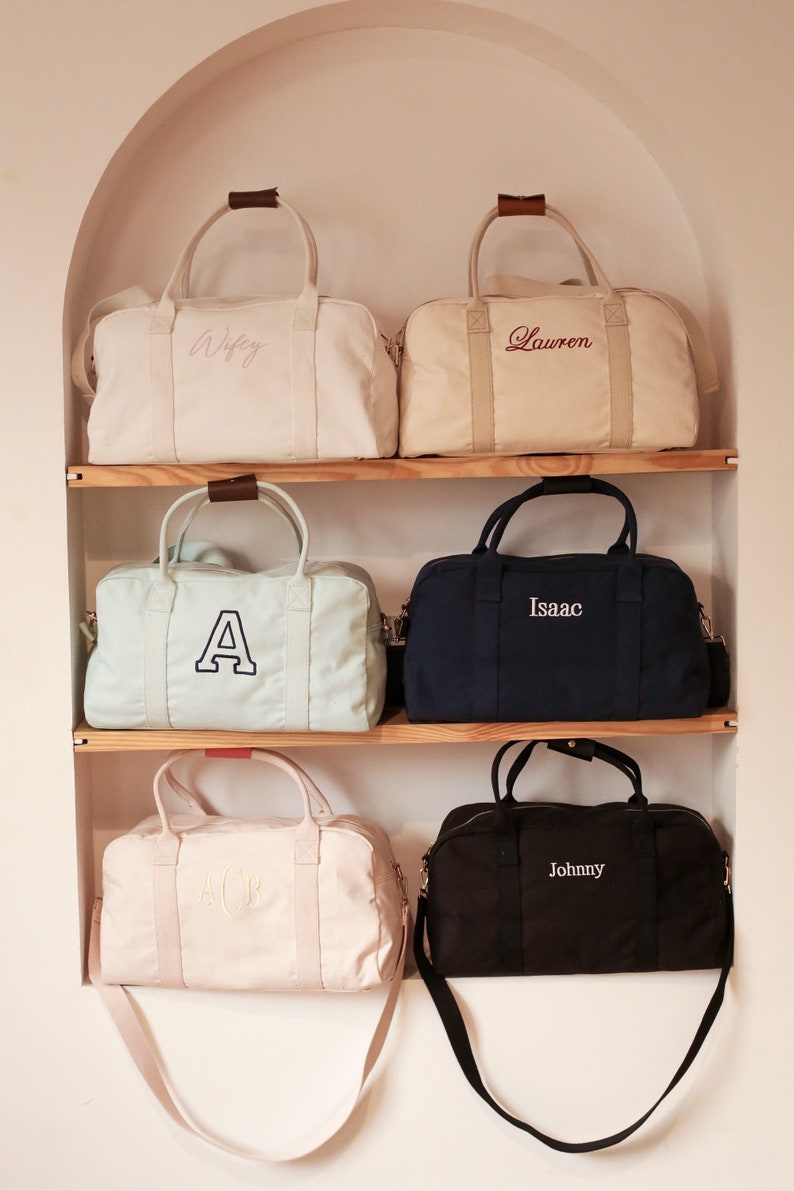 May include: Five canvas duffel bags with personalized embroidery on a wooden shelf. The bags are in various colors, including beige, light blue, navy blue, and black. The embroidery includes the names "Wifey", "Lauren", "Isaac", "ACB", and "Johnny".