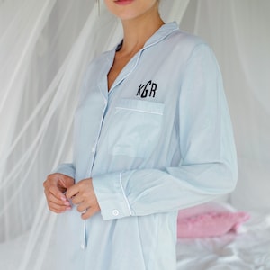 May include: A light blue button-down shirt with white piping and a black embroidered monogram "KGR" on the chest pocket.