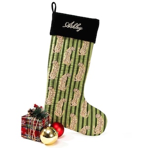 May include: A Christmas stocking with a green and tan leopard print pattern and black top. The name "Ashley" is embroidered in white script on the black cuff. A small gift box, red and gold ornaments complete the festive scene.