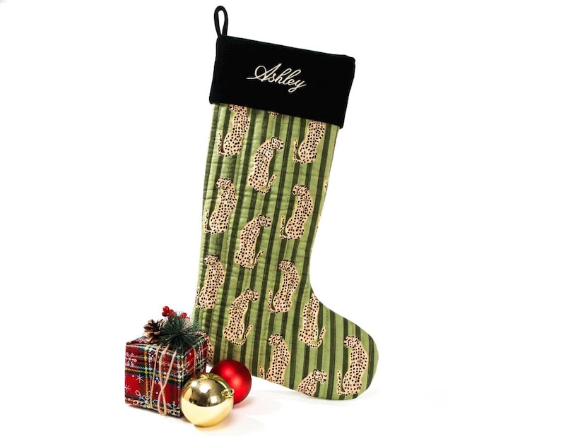 May include: A Christmas stocking with a green and tan leopard print pattern and black top. The name "Ashley" is embroidered in white script on the black cuff. A small gift box, red and gold ornaments complete the festive scene.