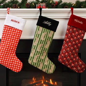 May include: Three Christmas stockings hang above a fireplace. One is red and white checkered, another green with leopard print, and the third is red with a floral pattern. Each stocking has a name embroidered on the cuff.