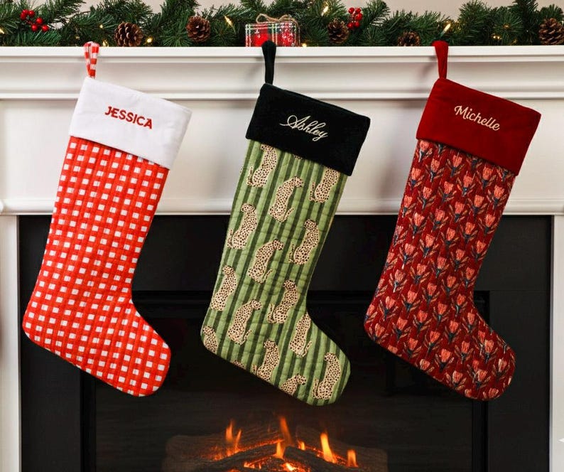 May include: Three Christmas stockings hang above a fireplace. One is red and white checkered, another green with leopard print, and the third is red with a floral pattern. Each stocking has a name embroidered on the cuff.
