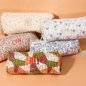 May include: A collection of quilted pencil cases in various floral patterns and colors. Some cases feature embroidered initials or names in red thread. Each case has a zipper closure and a tassel.