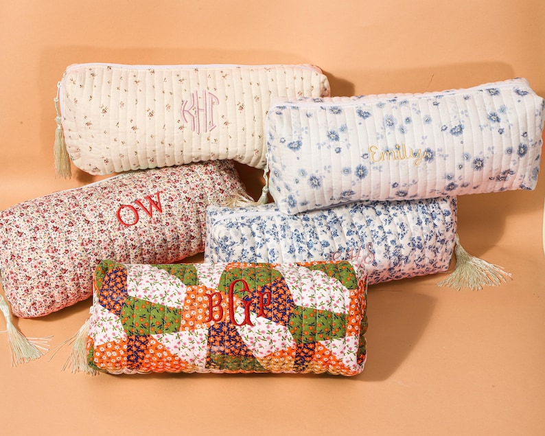 May include: A collection of quilted pencil cases in various floral patterns and colors. Some cases feature embroidered initials or names in red thread. Each case has a zipper closure and a tassel.