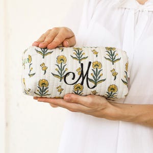 May include: A rectangular, quilted cosmetic bag with a floral pattern in yellow and green on a cream background. The bag features a large, embroidered black letter "M". The bag is held by a person wearing a white shirt.