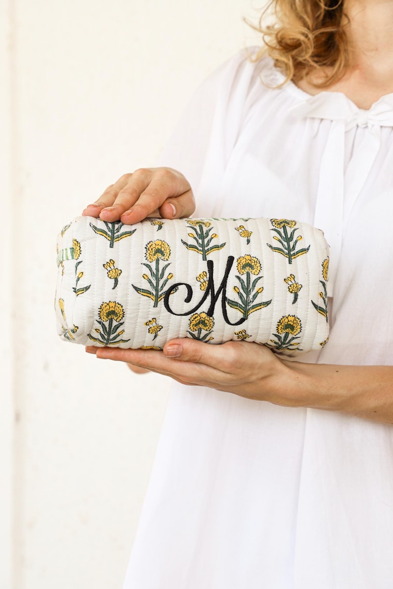 May include: A rectangular, quilted cosmetic bag with a floral pattern in yellow and green on a cream background. The bag features a large, embroidered black letter "M". The bag is held by a person wearing a white shirt.