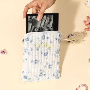 Puede incluir: Una funda blanca acolchada para lector de libros electr&oacute;nicos con un estampado floral azul y el nombre "Emily" bordado en amarillo. La funda est&aacute; abierta, sosteniendo un lector de libros electr&oacute;nicos. La imagen est&aacute; tomada desde arriba.