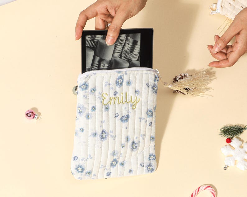 Puede incluir: Una funda blanca acolchada para lector de libros electr&oacute;nicos con un estampado floral azul y el nombre "Emily" bordado en amarillo. La funda est&aacute; abierta, sosteniendo un lector de libros electr&oacute;nicos. La imagen est&aacute; tomada desde arriba.