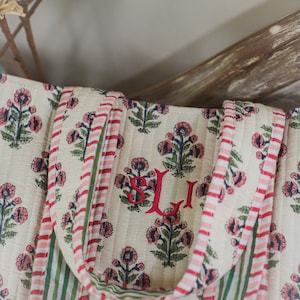Puede incluir: Bolso tote acolchado color crema con un estampado floral en rosa y azul marino. El bolso presenta asas y ribetes a rayas rojas y blancas. Las iniciales "SJI" est&aacute;n bordadas en rojo.