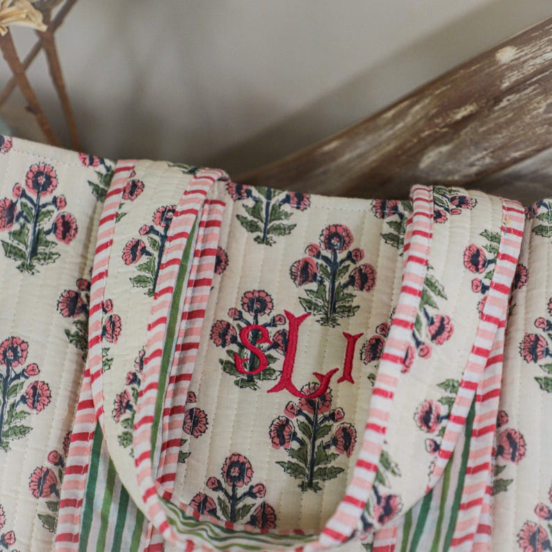 Puede incluir: Bolso tote acolchado color crema con un estampado floral en rosa y azul marino. El bolso presenta asas y ribetes a rayas rojas y blancas. Las iniciales "SJI" est&aacute;n bordadas en rojo.