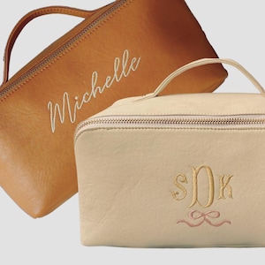 May include: Two zippered cosmetic bags, one brown with the name "Michelle" embroidered in white, and the other beige with the monogram "S.D.K." embroidered in gold with a pink ribbon.