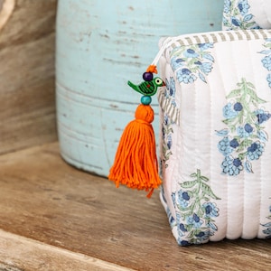 May include: A white quilted cosmetic bag with a blue floral pattern and a striped border. The bag features an orange tassel with a decorative bird charm. The background includes a light blue vase and a wooden surface.