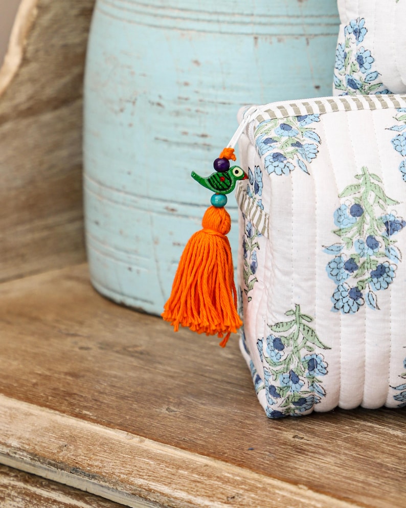 May include: A white quilted cosmetic bag with a blue floral pattern and a striped border. The bag features an orange tassel with a decorative bird charm. The background includes a light blue vase and a wooden surface.