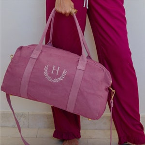 May include: A dusty rose canvas duffel bag with a gold zipper and a gold strap. The bag has an embroidered wreath with the letter "H" in the center.