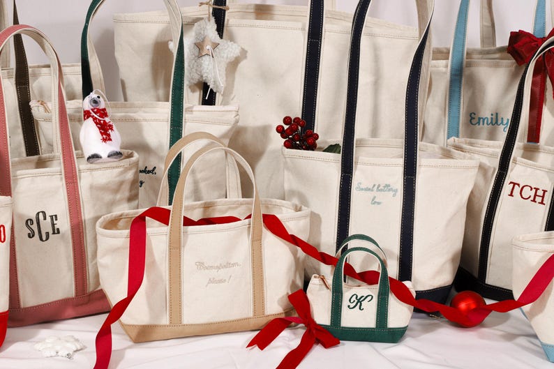 May include: A collection of canvas tote bags in various sizes and colors. Some bags have personalized initials or names embroidered on them. Red ribbons and decorative accents add a festive touch. The bags have different colored straps and trim.