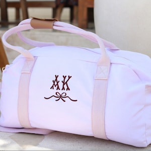 May include: A light pink canvas duffel bag with pink straps and a brown leather handle. The bag has a monogram of "MKK" with a bow embroidered on the front.