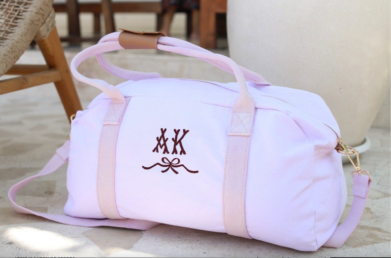 May include: A light pink canvas duffel bag with pink straps and a brown leather handle. The bag has a monogram of "MKK" with a bow embroidered on the front.