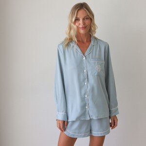 May include: A light blue satin pajama set with white piping and a monogrammed pocket. The set includes a long-sleeved shirt and shorts.