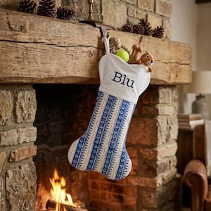 May include: A bone-shaped Christmas stocking featuring a white cuff embroidered with the name Blu in dark blue thread. The body of the stocking has a blue and white vertical striped pattern. It is filled with a tennis ball and a plush toy.