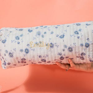 May include: A white quilted cosmetic bag with a blue floral pattern and the name "Emily" embroidered in gold. The bag has a tassel zipper pull and is held against a peach background.