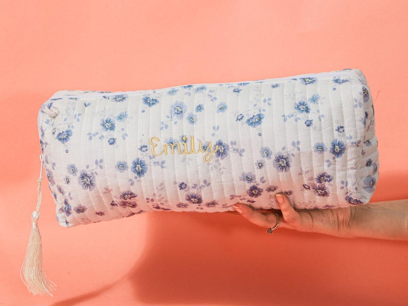 May include: A white quilted cosmetic bag with a blue floral pattern and the name "Emily" embroidered in gold. The bag has a tassel zipper pull and is held against a peach background.