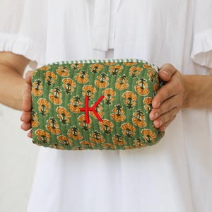 May include: A green quilted cosmetic bag with a floral pattern in yellow and orange. The bag has a red embroidered "K" and a green and white striped trim. The bag is held by a person wearing a white dress.